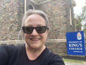 Woman stands in front of old stone building with sign in background saying "University of King's College"
